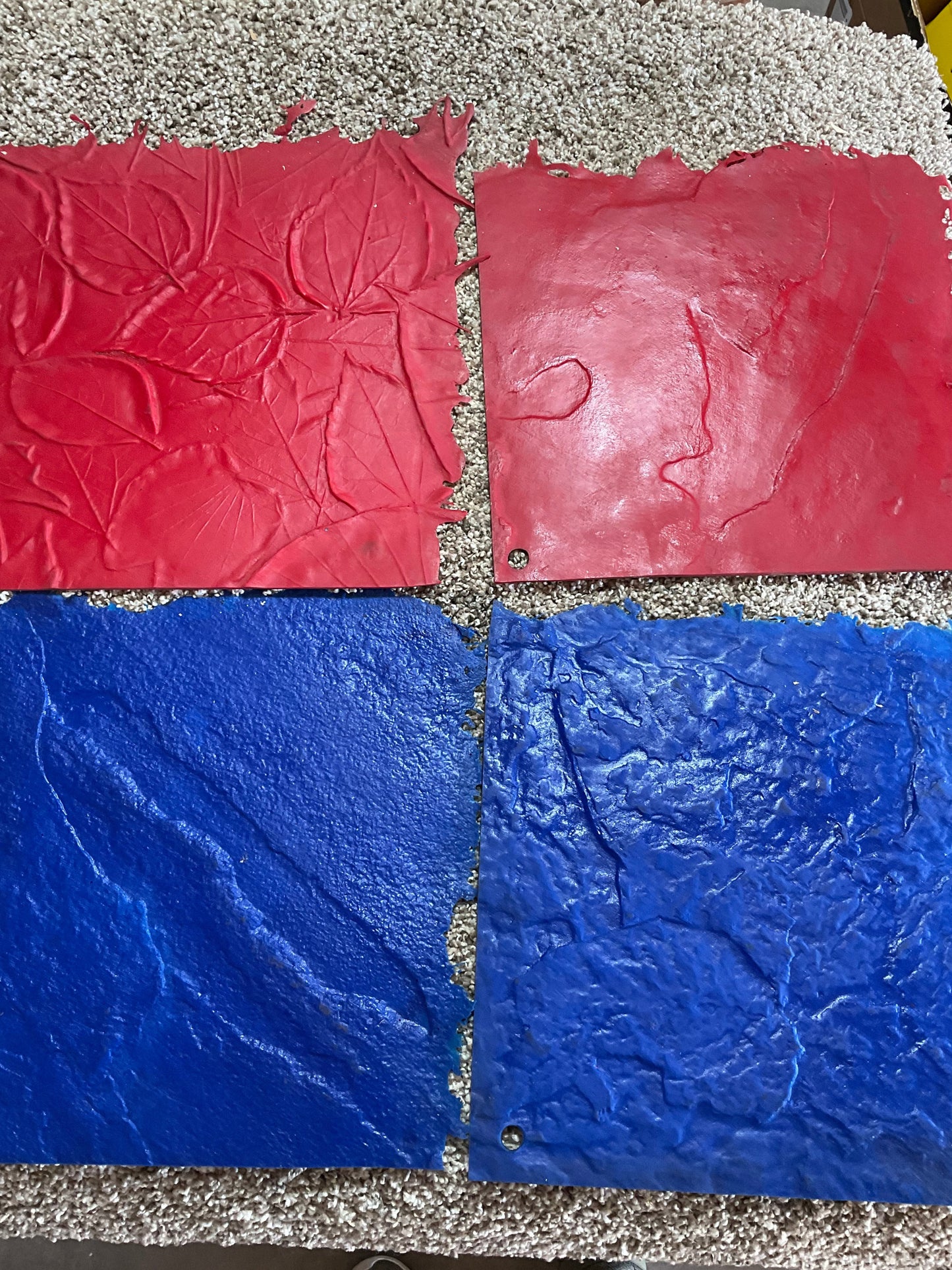 four square landscape curbing stamp texture mats displayed on top of a rug. The top two are red, with the left one featuring a leaf pattern and the right one having a smoother texture with some irregular marks. the bottom two are blue, both showcasing natural stone textures with visible veins and cracks. the edges of the stamps are slightly rough, and the image is taken from a top-down perspective.