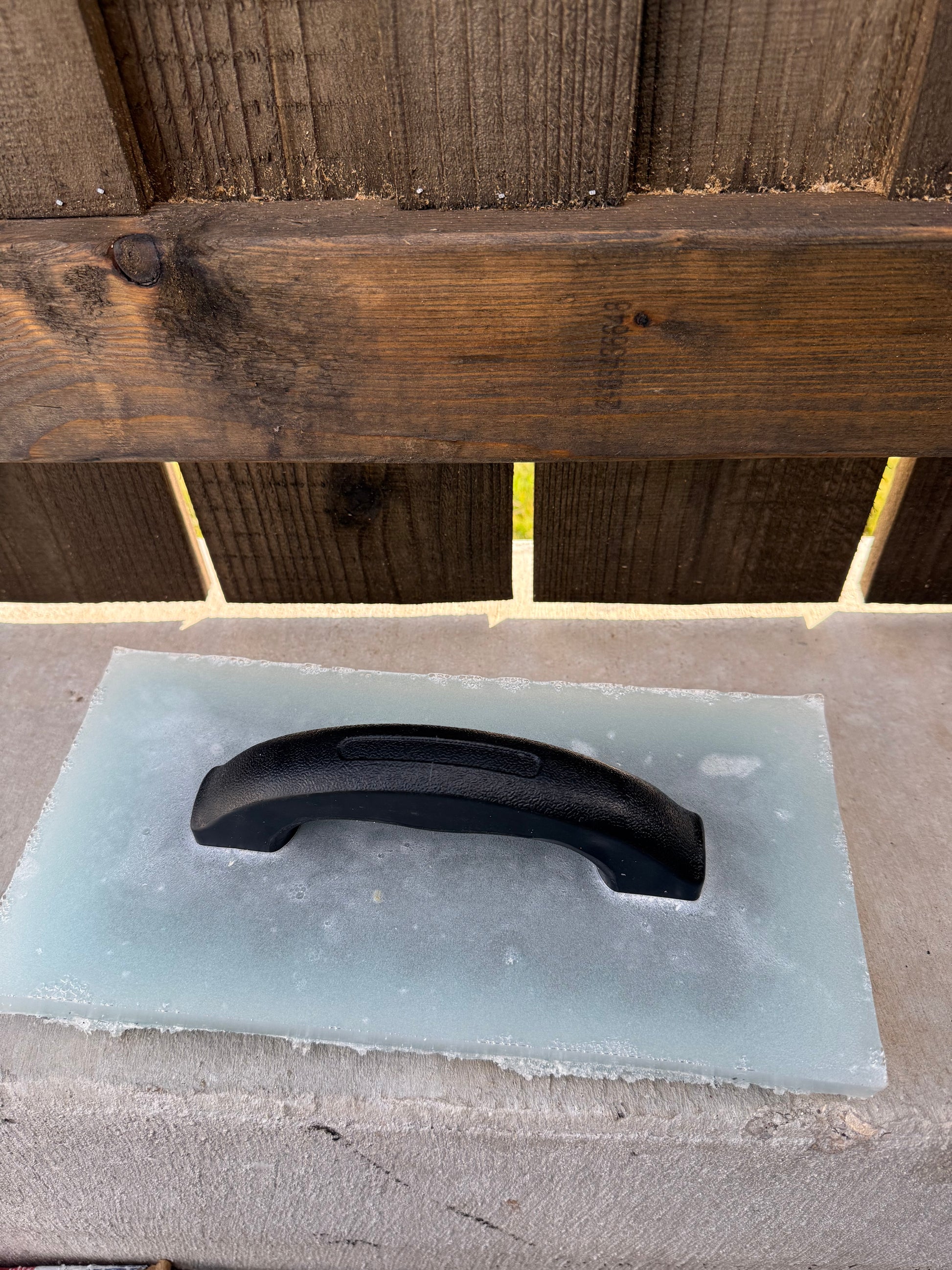 Black handle on the gabbro textured trowel on a concrete wall next to a wooden fence.