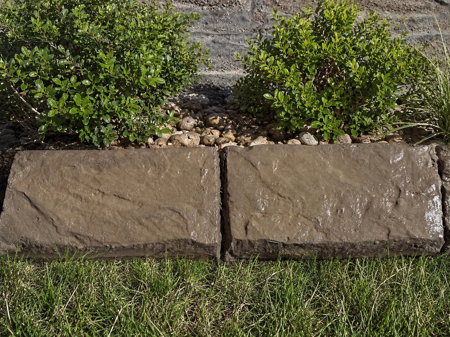 Two rectangular stone border pavers in a natural brown-gray textured finish, shown installed in a garden bed with grass, small rocks, and green shrubs.