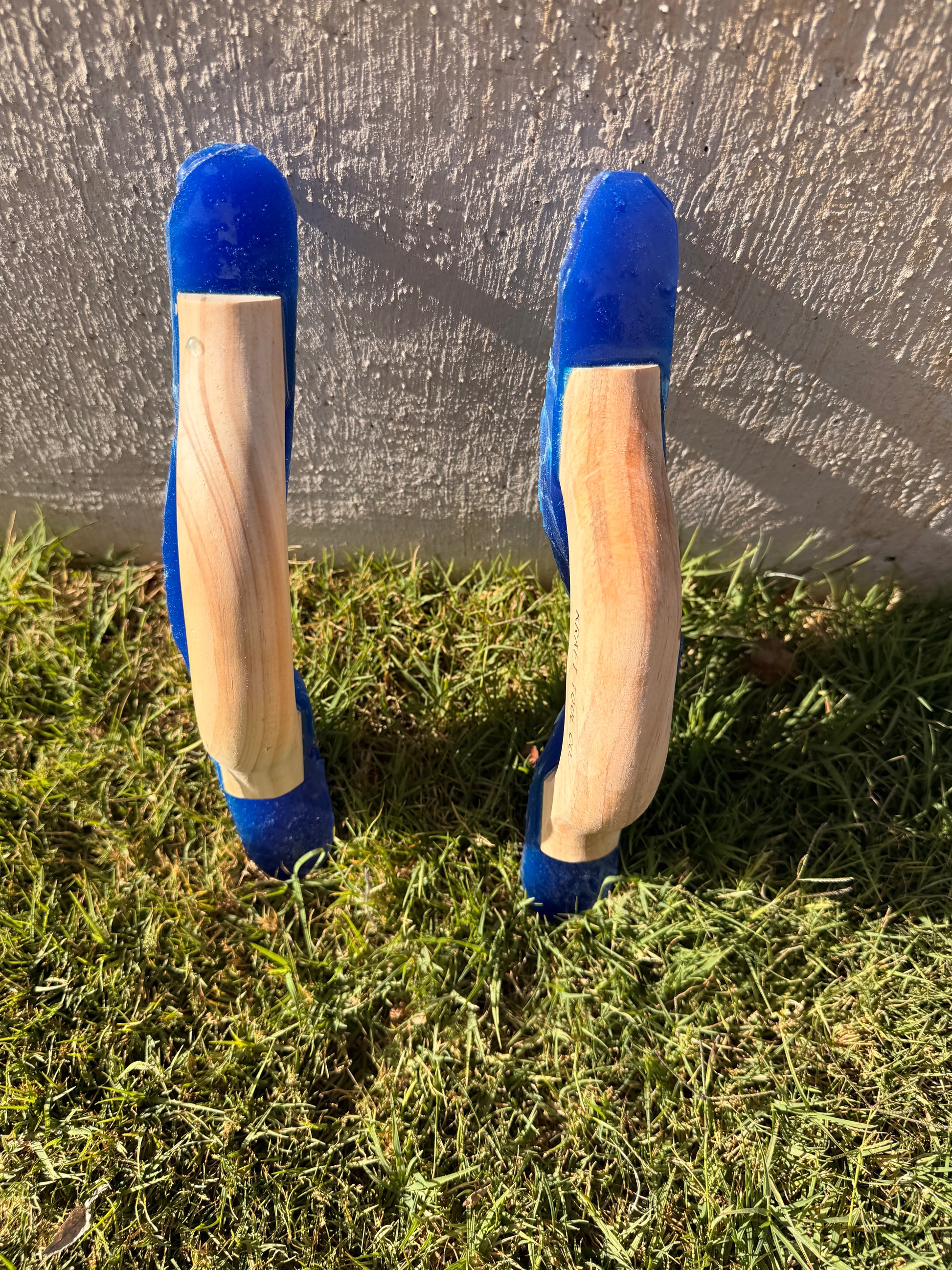 Two wooden joint tools with blue tops on grass against a concrete wall.